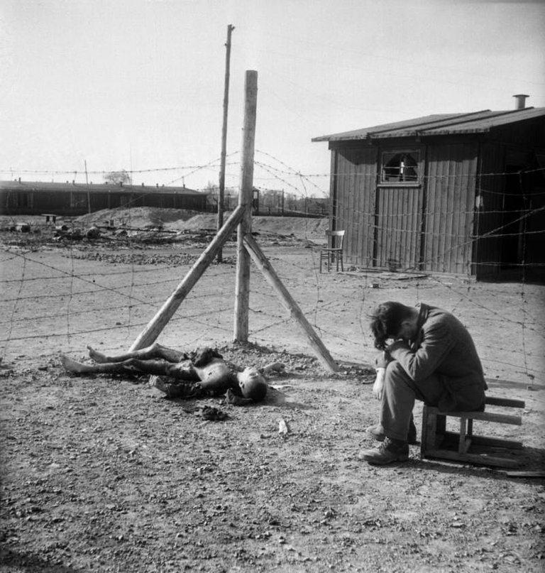 A young man sits on an overturned stool next to a burnt body inside the Thekla concentration subcamp outside Leipzig, Germany soon after its liberation by U.S. forces on April 18, 1945..jpg